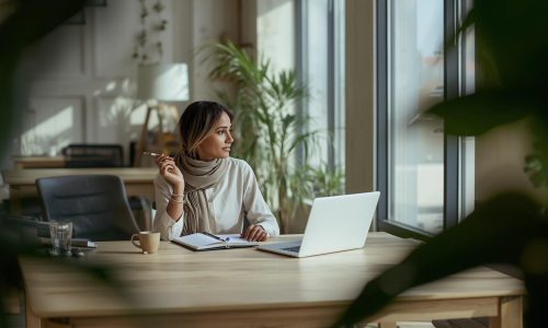 Solopreneure assise sur une chaise de bureau devant son laptop dans une position pensive. En pleine réflexion pour le développement de son activité entrepreneureuriale. Ambiance open-space calme, végétalisé, sobre et professionnelle